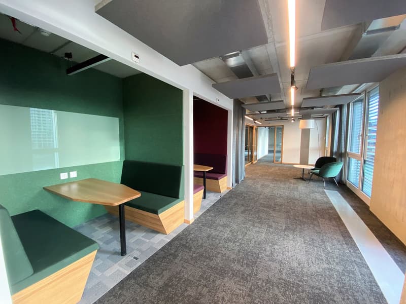 A corridor with carpet flooring, green benches, wooden tables, white walls, ceiling lights, and windows.