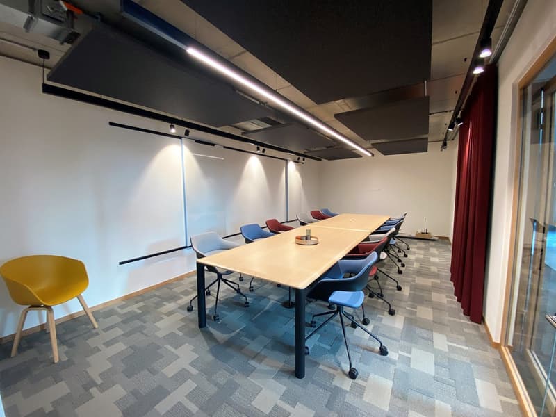 meeting room with a white wall, a wooden table, several blue, red, and yellow chairs, a yellow chair on the left, and a red curtain at the end of the room.