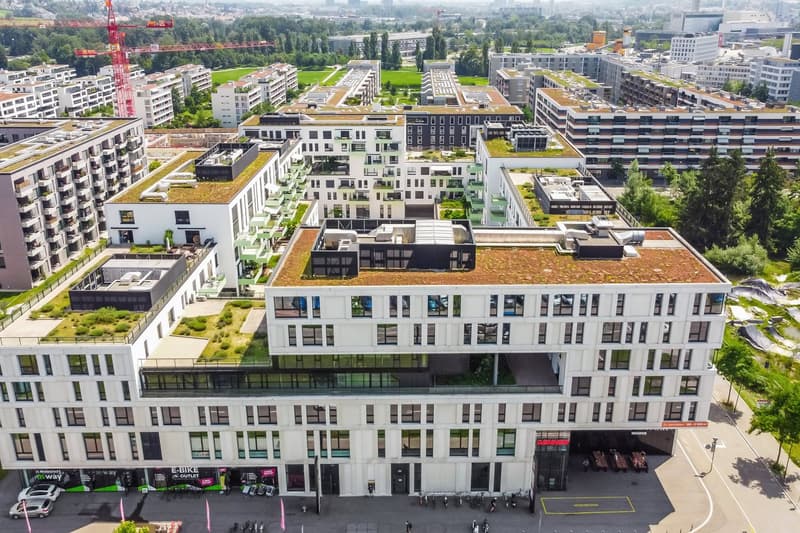 High-rise building with green roofs, glass windows, multiple balconies, ground-level stores, and parking area