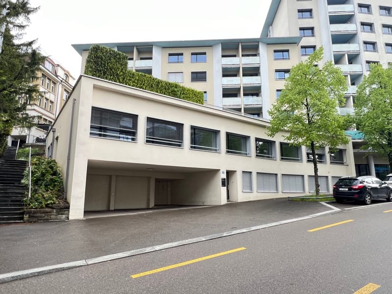 An apartment building with multiple windows and balconies. There is a garage entrance at the front of the building, and the parking lot is to the left of the building. The building is next to a building with trees and staircases.