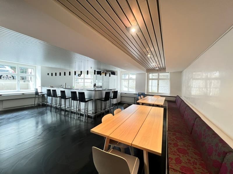 A room with a wooden table, chairs, a long bench, a bar counter, and windows