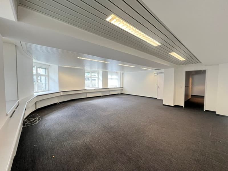 Empty room with white walls, black floor, three windows, and a ceiling with lights