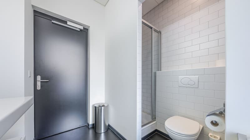 Bathroom with white tiles, glass shower, toilet, trash bin, and toilet paper holder