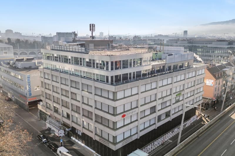 A large multi-story building with many windows, located in an urban area with other buildings and a road in front.