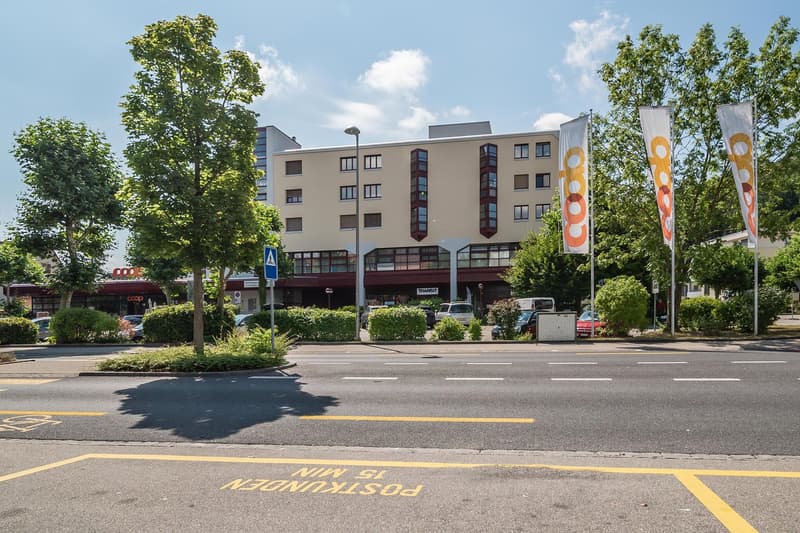 A multi-story commercial building with a retail space on the ground floor, surrounded by trees and a parking area. The building has a modern design with large windows and colorful flags or banners displayed.