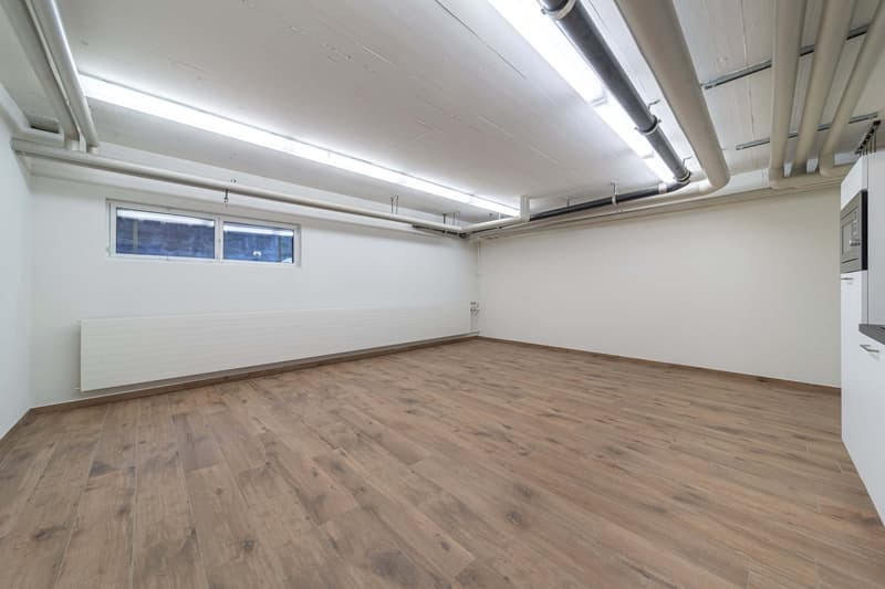 This image shows a large, open room with a high ceiling and exposed ductwork. The room has white walls and a wooden floor with a rustic, distressed appearance. There are two small windows near the ceiling, providing natural light. The room appears to be a