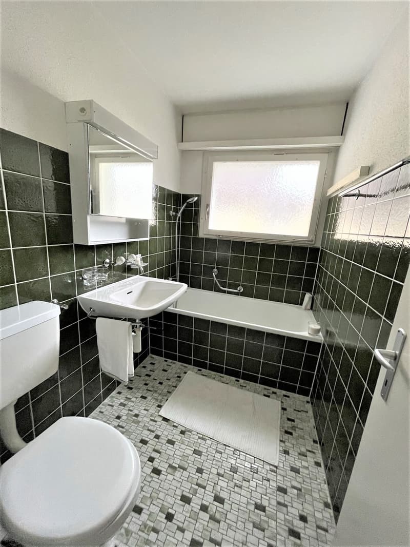 Bathroom with green tiles, white sink, white toilet, bathtub, mirror, towel rack, and a window