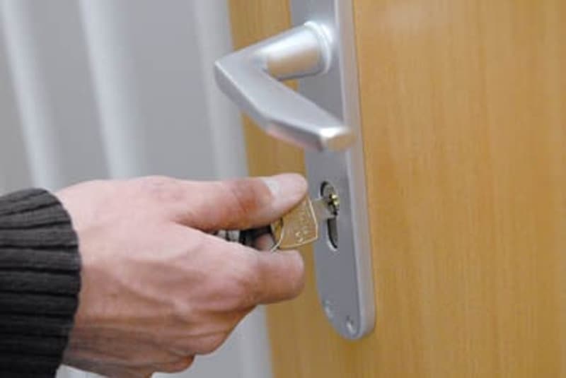 The image shows a person's hand unlocking a door handle. The door appears to be made of wood with a metal door handle and lock mechanism.