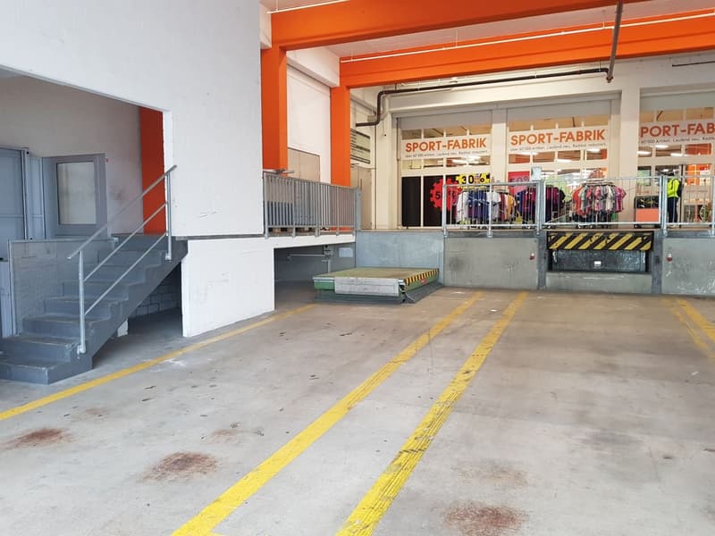 The image shows the interior of a sports retail store called 'Sport-Fabrik'. The store has an open layout with shelves and racks displaying various sports clothing and equipment. The walls are painted in an orange color, and there is a garage-like entranc