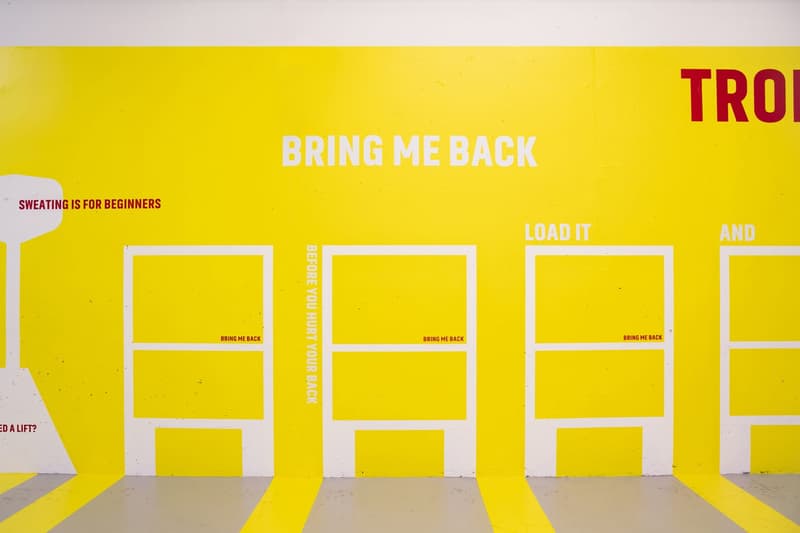 The image shows a yellow wall with various text and graphics. The main text says 'BRING ME BACK' in large letters. There are also frames or boxes with the text 'BRING ME BACK' inside them. The wall also has other text such as 'SWEATING IS FOR BEGINNERS',
