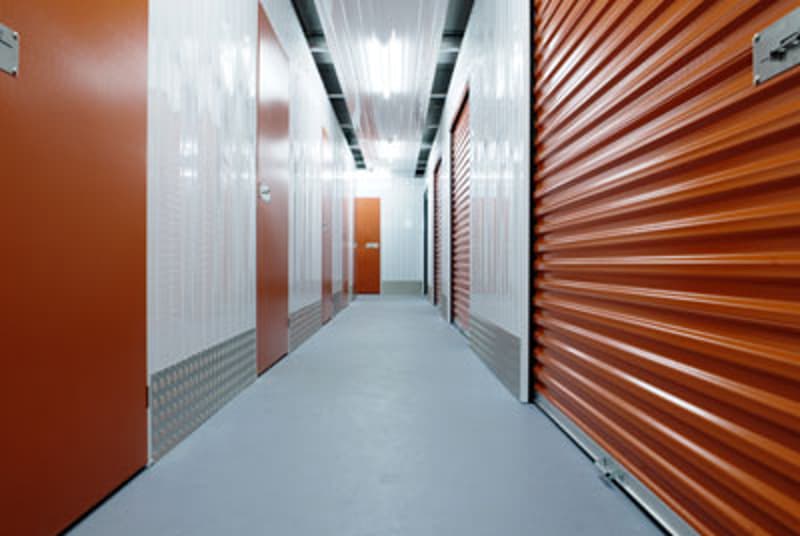 The image shows a long hallway with rows of orange storage units on both sides. The hallway has a concrete floor and the walls are made of white and orange panels. The lighting is bright, creating a clean and organized atmosphere typical of a self-storage