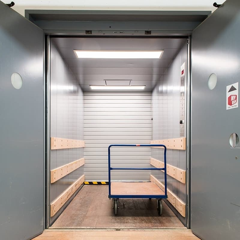 The image shows the interior of an elevator. The elevator has a metal frame and shelves on the sides, likely for storing items. There is a blue cart or trolley inside the elevator, which appears to be used for transporting goods or materials.