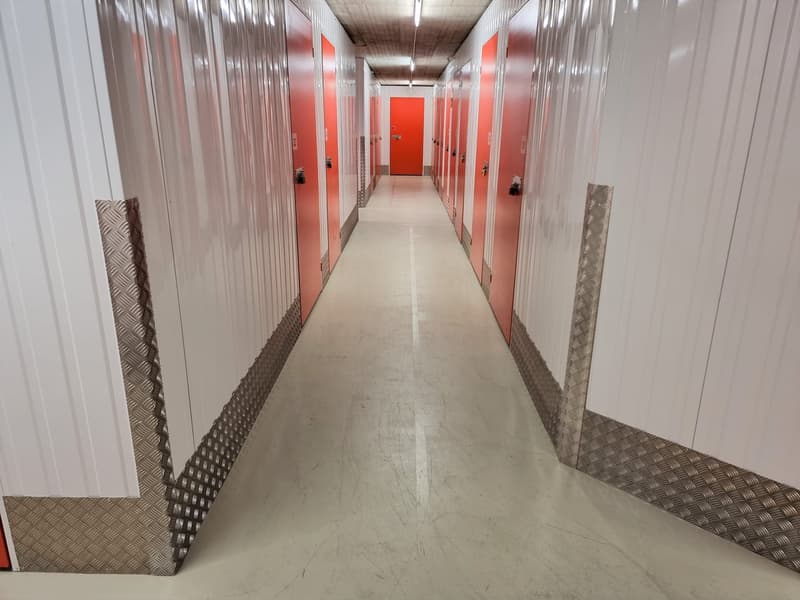 The image shows a long, narrow hallway with multiple storage units on both sides. The hallway has a concrete floor and the storage units have red doors. The walls are made of a corrugated material, likely for insulation or soundproofing. The overall appea