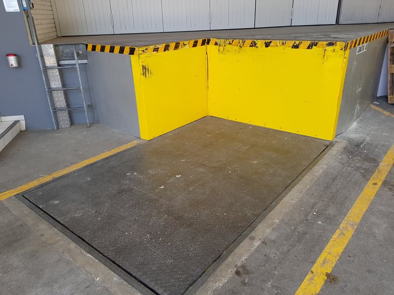 The image shows a large yellow garage door with black and yellow striped trim. The floor in front of the garage door is a black diamond-patterned surface, likely a rubber or metal material used for loading and unloading. There is a ladder visible on the s