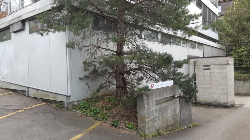 A multi-story concrete building with large windows, surrounded by trees and a concrete structure that appears to be a parking area or entrance.