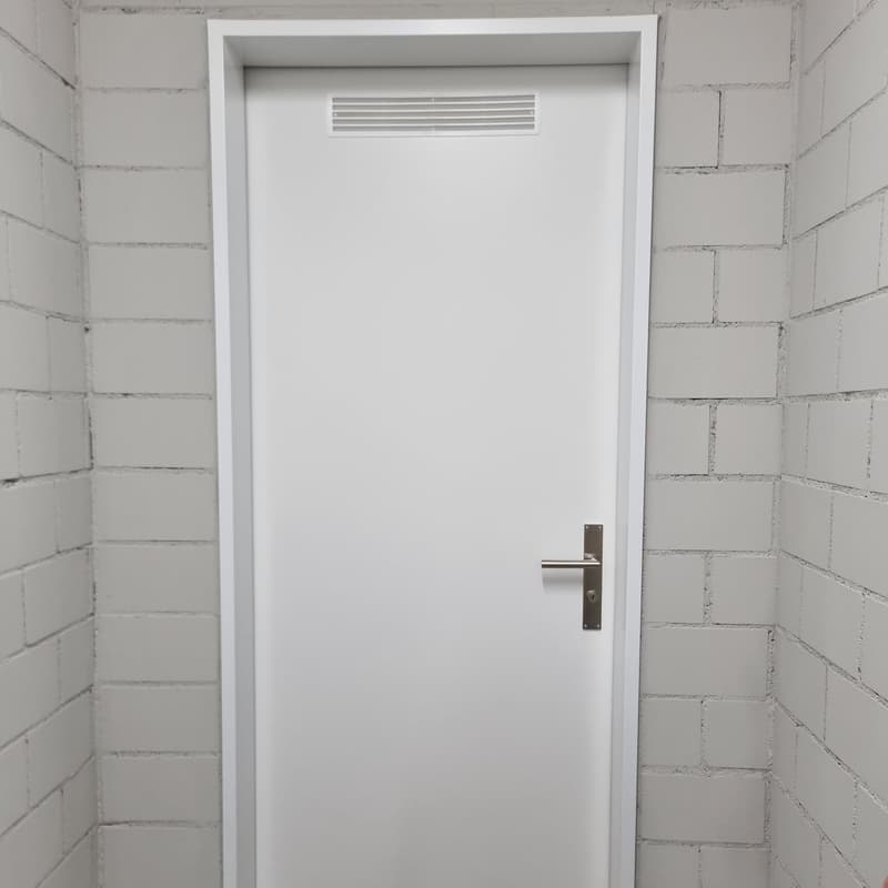 A closed white door with a metal handle and a white air vent above the door on a white brick wall.