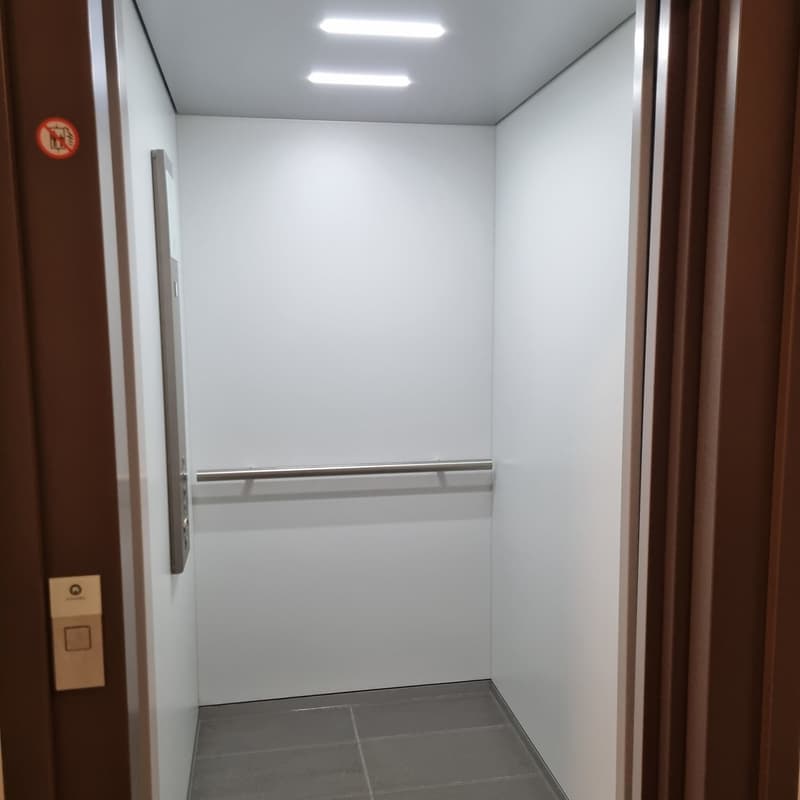 empty elevator with white walls and tiled floor, metallic handrail, lights on ceiling
