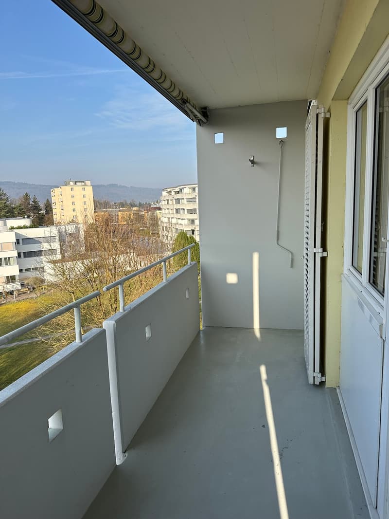 Empty balcony with railings, white walls, tiled floor, entrance from room, buildings in the background