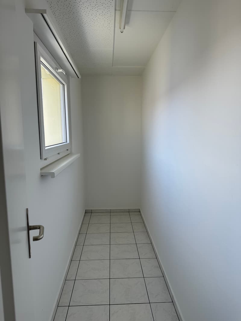 Empty hallway with white tiles, white walls, window with white frame, and white ceiling with perforations.