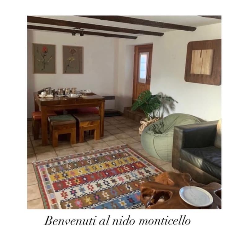 Rustic living room with exposed wooden beams, tile flooring, wooden dining table and chairs, artwork on the walls, and a bean bag chair. There is also a fireplace and what appears to be a washing machine visible in the room.