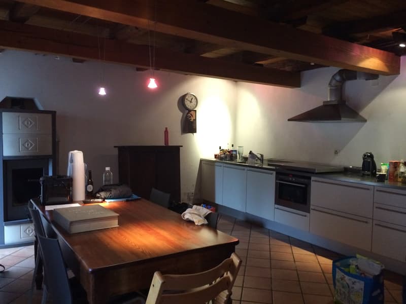 The image shows a kitchen with a wooden dining table, dark cabinets, a dishwasher, and a washing machine. The kitchen has a rustic, industrial style with exposed wooden beams on the ceiling and tile flooring.