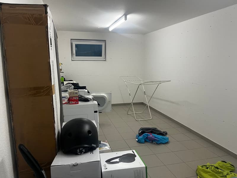 The image shows a utility room with a washing machine, a clothes drying rack, and various other household items and boxes. The room has white walls and a tiled floor.