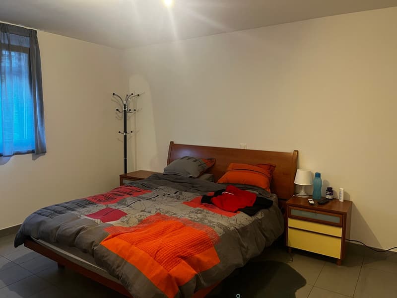 Bedroom with a wooden bed frame, bedside table, and a coat rack. The room has a messy bed with gray and orange bedding, and a window with curtains. There is also a dresser or cabinet in the room.