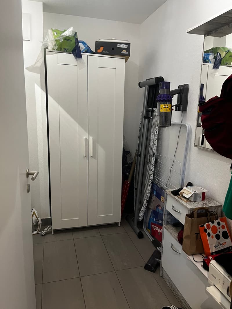 The image shows a small utility room or storage area. There are two white cabinets, a vacuum cleaner, and various boxes and items stored in the room. The floor is tiled.