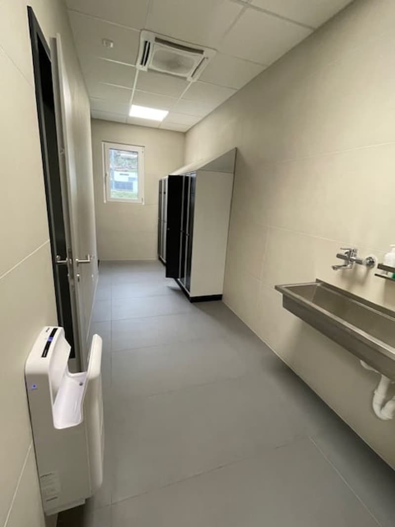 The image shows a hallway or corridor with a tiled floor, white walls, and a window. There is a hand dryer or air hand dryer mounted on the wall. The hallway leads to a room or space with a door and what appears to be a refrigerator or appliance visible t