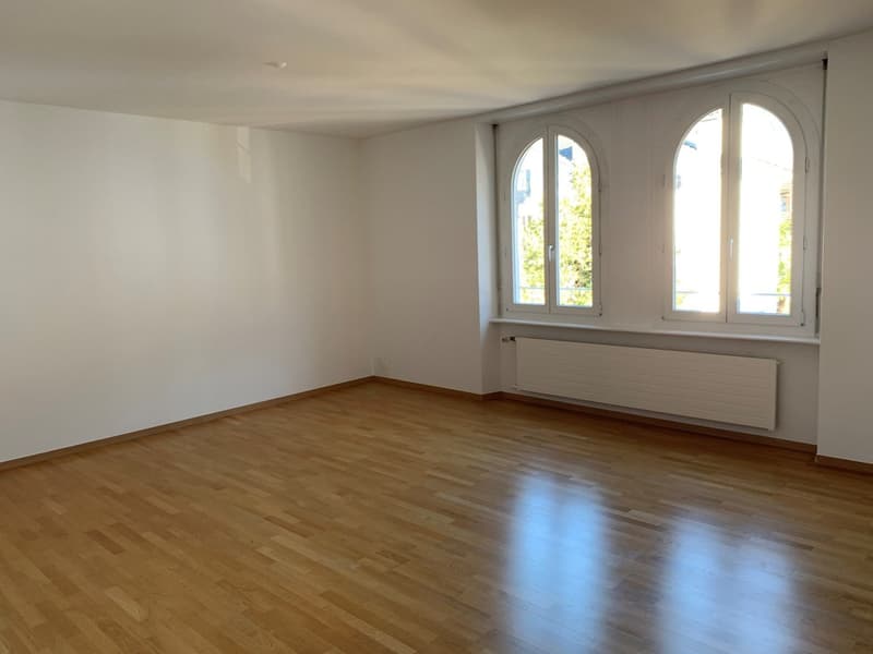 This image shows a spacious room with white walls and large arched windows that let in plenty of natural light. The room has a hardwood floor that appears to be in good condition. There is a radiator visible along one of the walls, suggesting this is like