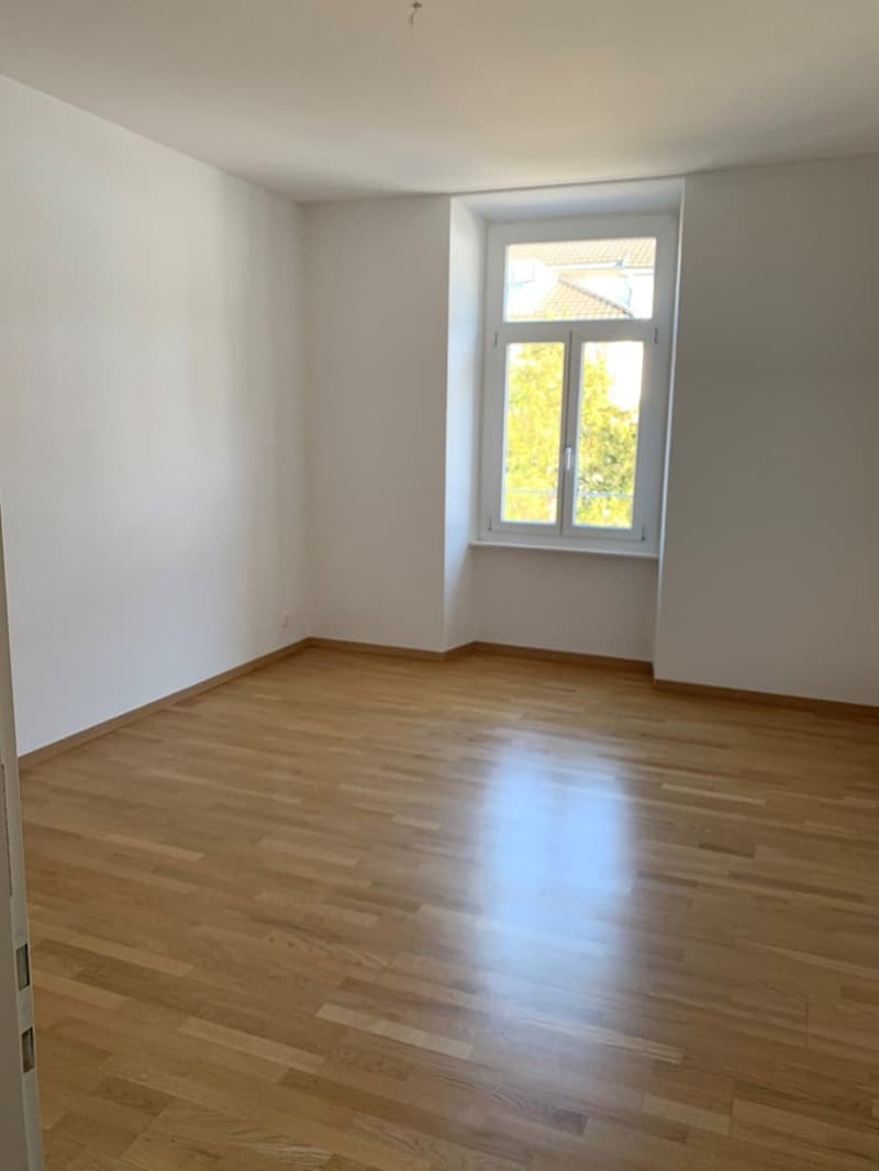 This image shows an empty room with white walls and a large window overlooking the outdoors. The room has a hardwood floor that appears to be newly installed or refinished, with a glossy finish. The room appears to be a living room or bedroom, with no vis