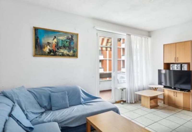 Living room with white tiles, white walls, blue sofa, coffee table, wooden entertainment center, television, wooden balcony doors, framed artwork, white curtains.
