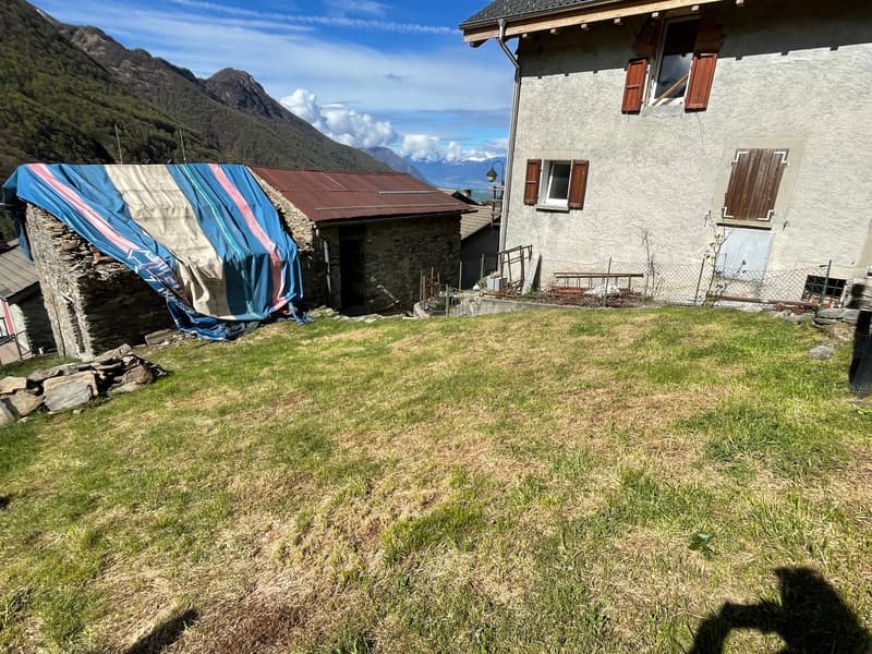 Two-story stone house with wooden shutters, surrounded by a grassy yard and mountains in the background. There is a blue and pink tarp covering an outdoor structure or storage area.