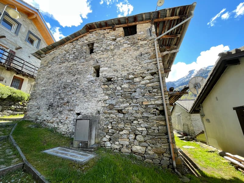 Stone building, exposed stone walls, metal roof, a door with a box in front of it, metal pipe leading to the roof, grass, mountains in the background.