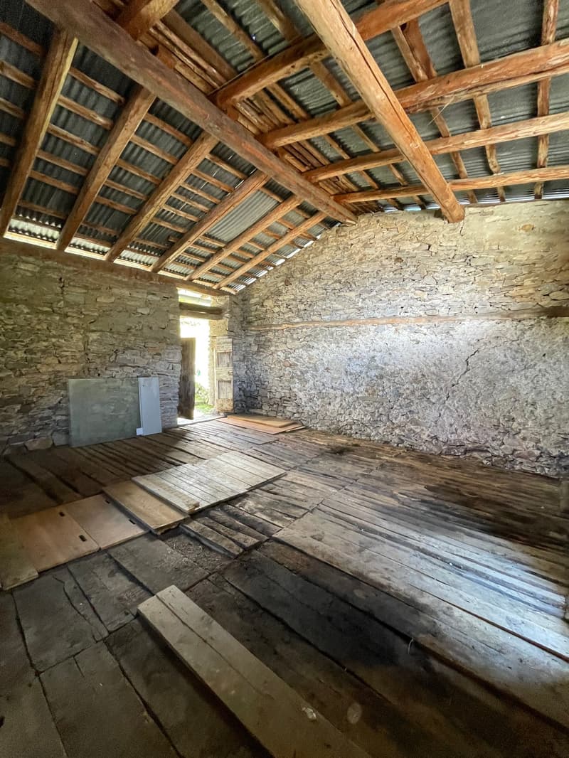 The image shows the interior of an old, rustic building. The walls are made of stone, and the ceiling is constructed with wooden beams and a corrugated metal roof. The floor is made of wooden planks. The space appears to be a storage or utility area, with