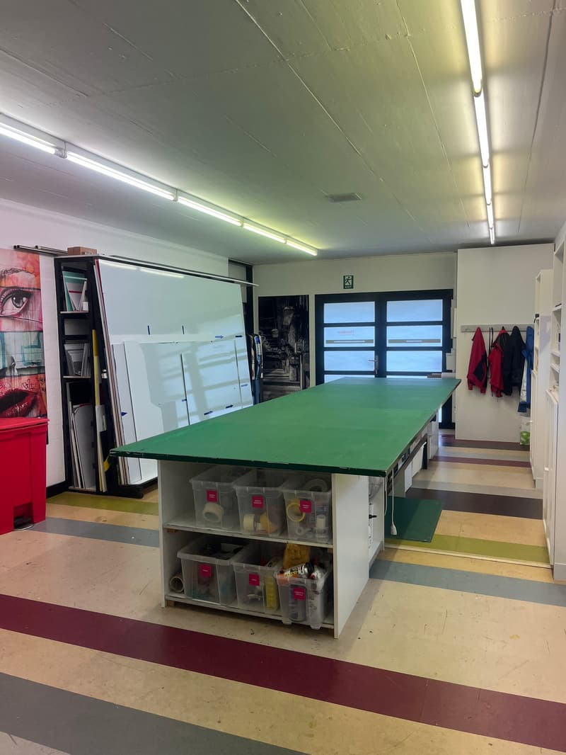 A large, rectangular, green table, surrounded by cabinets filled with various items. In the background, there is a wall with a whiteboard and a tall bookshelf full of artwork. To the right, there is a large door, with three coats hanging near it.