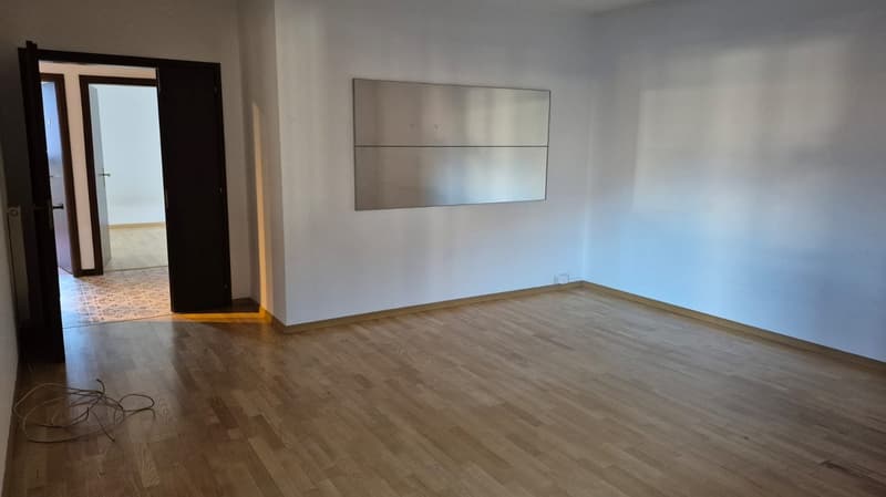 Empty room, wooden floor, white walls, open door to hallway, large mirror on wall