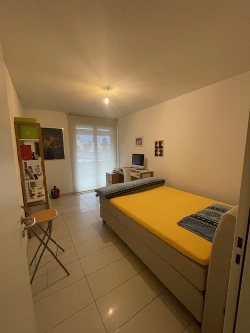 Single bedroom with a bed, desk, monitor, books on a shelf, and a chair