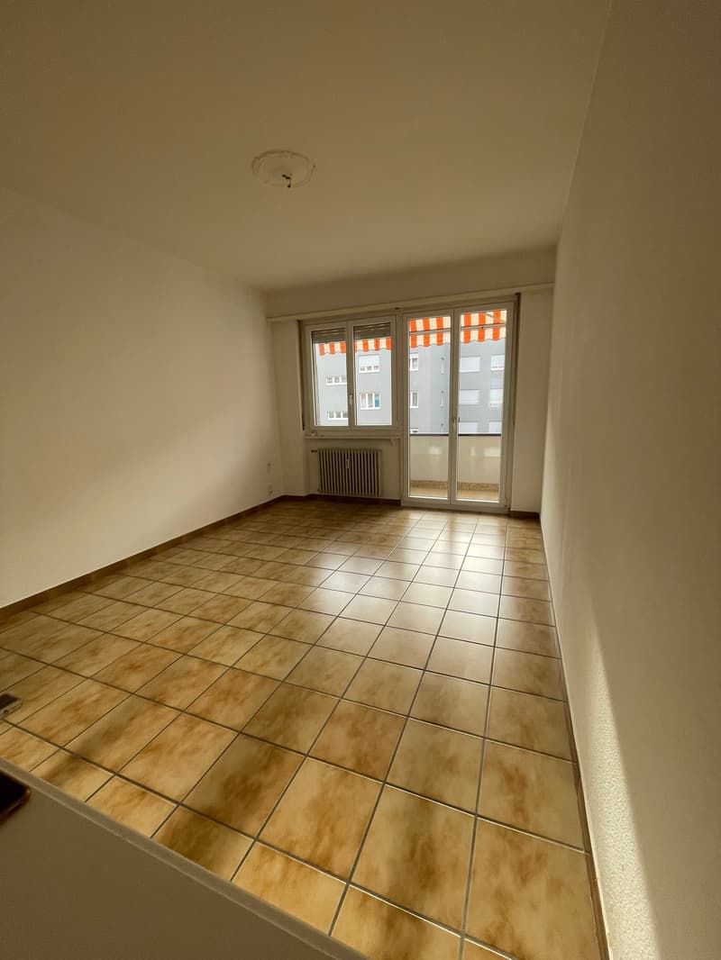 Empty room, light brown tiles, glass sliding doors to balcony with awning, heater mounted on wall, power outlet on wall