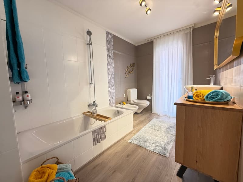 Bathroom with bathtub, shower, toilet, sink, wooden floor, white tiles, towels, and toiletries