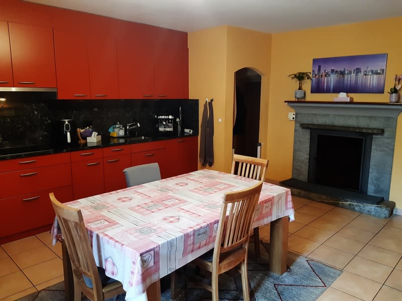 The image shows a combined kitchen and dining area. The kitchen features red cabinets and a black countertop. There is a dining table with a patterned tablecloth and wooden chairs. A fireplace is visible in the corner of the room, and a large cityscape ar