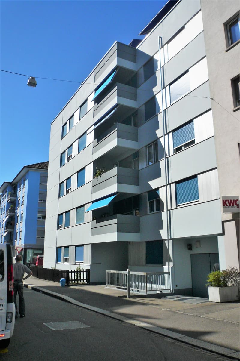 Modern apartment building with multiple balconies, white exterior, blue awnings, and a fenced entrance.