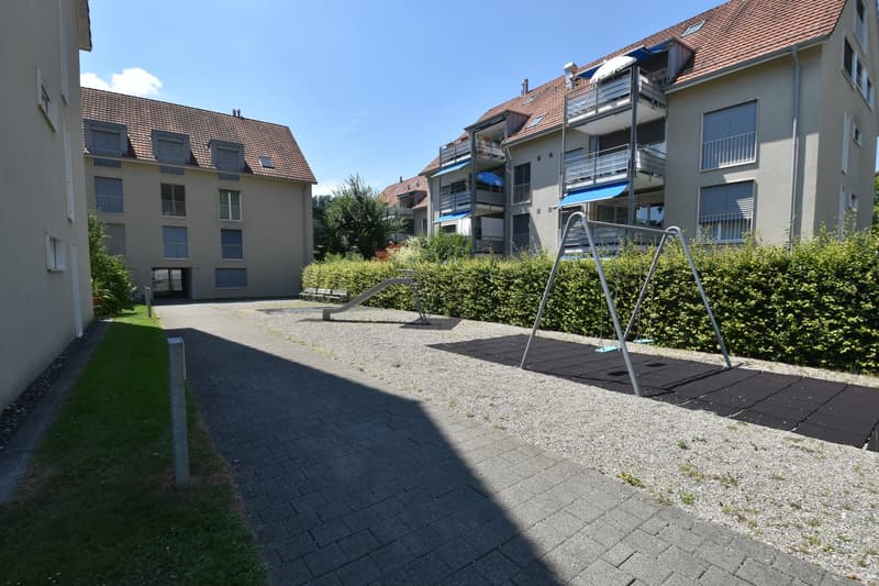Two buildings with balconies, brick walls, and red roofs. A paved area with a swing set and slide. Green hedges and a grassy area.