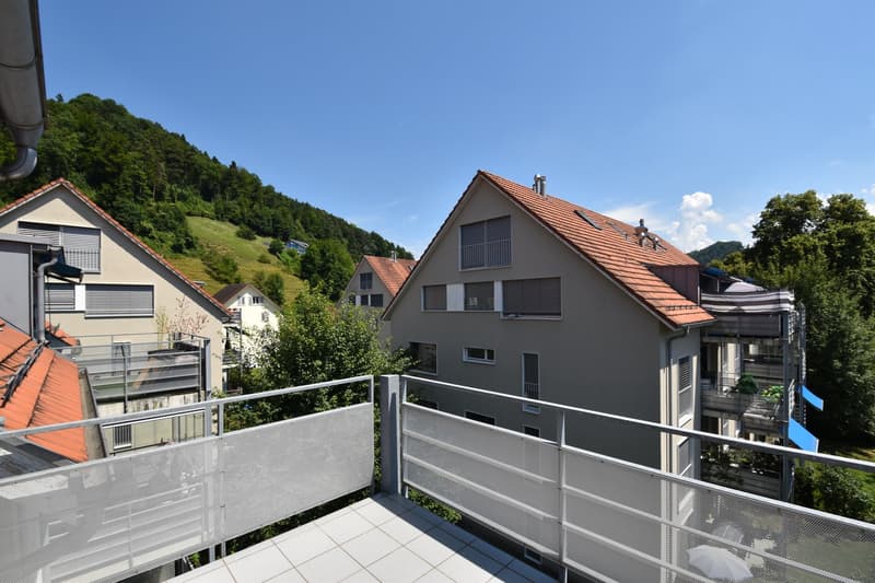 The balcony has a railing and is surrounded by a metal fence. The house has a brown roof, and glass windows. The balcony overlooks a mountain and houses in the background.
