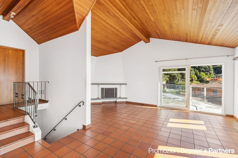 Spacious living room with high vaulted wooden ceiling, white walls, terracotta tile flooring, and a fireplace. Large windows provide a view of the outdoor area. The room has an open layout with a staircase leading to another level.