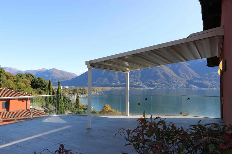 Covered outdoor terrace with a panoramic view of a lake surrounded by mountains. The terrace has a tiled floor and is furnished with potted plants. The view includes a body of water, mountains, and a clear blue sky.