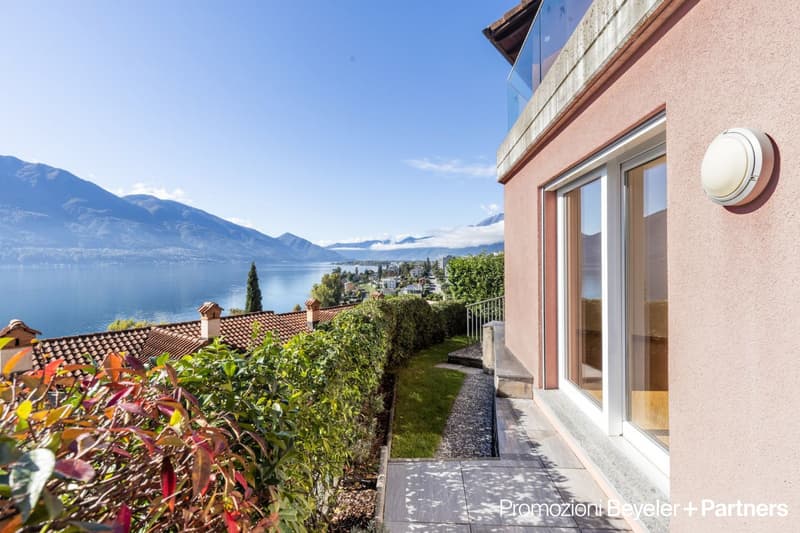Two-story house with a tiled roof, surrounded by lush greenery and overlooking a scenic lake and mountains in the background. The house has large windows and a balcony or terrace offering a panoramic view of the beautiful natural landscape.