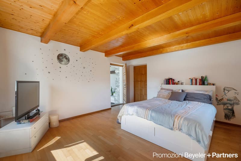 bedroom with wooden floor, white walls, wooden beams, flat screen TV, white cabinet, bed, pillow, blanket, wall decorations, wooden door, open door with view to outside