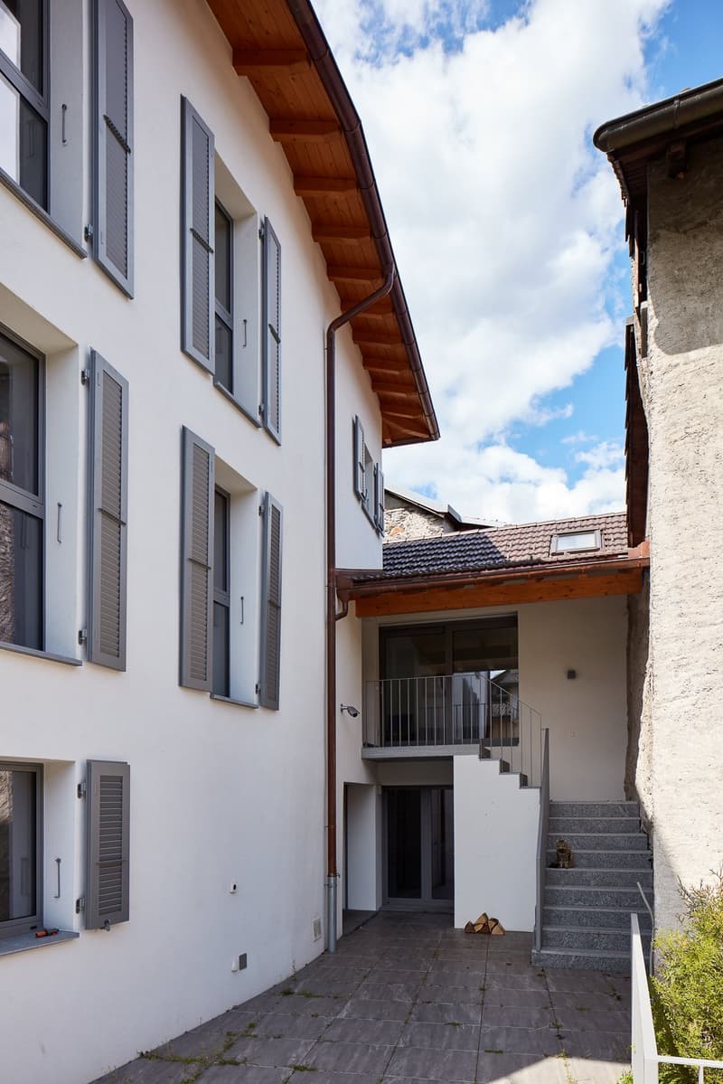 white building, multiple windows, wooden roof, stairs leading to entrance, paved courtyard