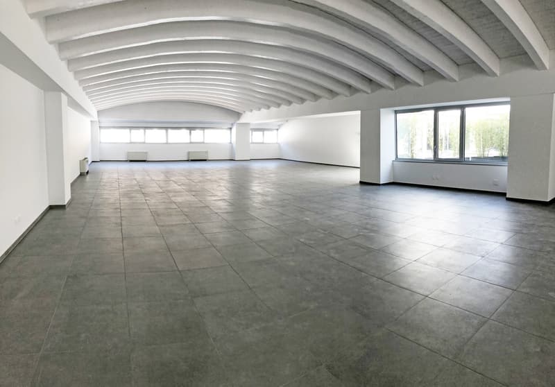 Large open-plan office space with high ceilings, exposed beams, and large windows providing natural light. The space has a minimalist design with white walls and gray tile flooring. There are several seating areas and a spacious layout suitable for variou
